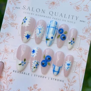 Op de afbeelding: Een set salon-kwaliteit press-on nagels met diverse ontwerpen. De nagels hebben een lichtroze basis met witte en blauwe accenten, waaronder bloempatronen, ruitjes en blauwe bessen. De tekst "REUSABLE | STURDY | STRONG" is zichtbaar.