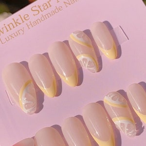May include: A set of ten false nails with a light pink base and a yellow lemon design. The nails are shaped like almonds and have a white outline around the lemon design. The nails are displayed on a pink card with the text "Twinkle Star Nails Luxury Handmade Nails" at the top.