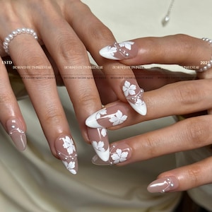 May include: Almond-shaped artificial nails with a white floral design and pearl accents. The nails have a nude base with white tips and white flower patterns. The text "DESIGNED BY TWINKLESTAR" is visible.