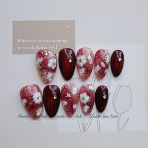 May include: A set of ten almond-shaped press-on nails featuring a deep red and pink color palette. The design includes hand-painted white cherry blossoms, gold leaf accents, and small clear rhinestones. The text reads Twinkle Star Nails.