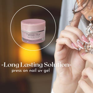 Puede incluir: Un frasco rosa de gel UV para uñas postizas Twinkle Star. El frasco tiene una tapa rosa claro y una base transparente, con el nombre de la marca y los detalles del producto impresos. También se ve una mano de mujer con uñas largas y manicuradas.