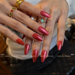 May include: Close-up of hands with long, red glitter nail polish. The nails are almond-shaped. The hands are adorned with gold rings, including a large ring with a colorful gemstone and a ring with a pearl.