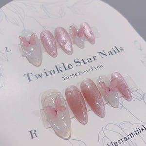 以下が含まれることがあります： Twinkle Star Nailsと書かれた台紙に飾られた10枚のネイルチップ。ピンクのキャッツアイ仕上げで、小さな白いパールをあしらった透明な部分と、親指と薬指のネイルには半透明のピンクの蝶のアクセントが施されています。