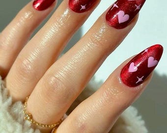 May include: Almond-shaped nails featuring a deep metallic red base color. Each nail is decorated with small, light pink heart shapes and tiny four-pointed star accents scattered across the surface. The finish is glossy and reflective.