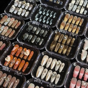 May include: A collection of multiple clear plastic storage cases, each containing a set of press-on artificial nails. The nails feature various designs, including solid colors, glitter finishes, ombre gradients, and 3D embellishments like bows.