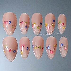 Peut inclure: Dix faux ongles ovales de couleur nude, chacun orné d'une guirlande de lumières de Noël colorées. Le texte "REUSABLE | STURDY | STRONG" est visible. Les ongles sont disposés sur une surface réfléchissante.