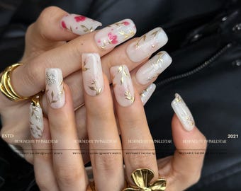 May include: A set of long, square-shaped artificial nails. Each nail is painted white with gold and silver floral designs, including leaves and small pink flowers. The text "DESIGNED BY TWINKLESTAR" and "REUSABLE SALON QUALITY NAILS" is visible.