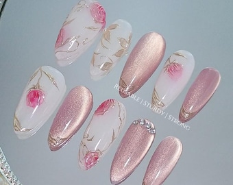 May include: A collection of ten artificial nails, each with a unique design. Some nails are rose gold, others are clear with pink roses and gold leaf details. One nail is decorated with rhinestones. The text "REUSABLE | STURDY | STRONG" is visible. The nails are displayed on a reflective surface.
