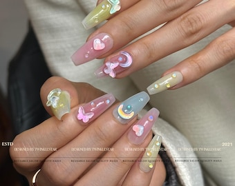 May include: A set of long, coffin-shaped acrylic nails with various 3D embellishments. Designs include butterflies, hearts, and crescent moons in pastel pink, yellow, and blue hues. The nails are decorated with iridescent glitter and small gems. Text on the image reads "ESTD", "DESIGNED BY TWINKLESTAR", and "2021".