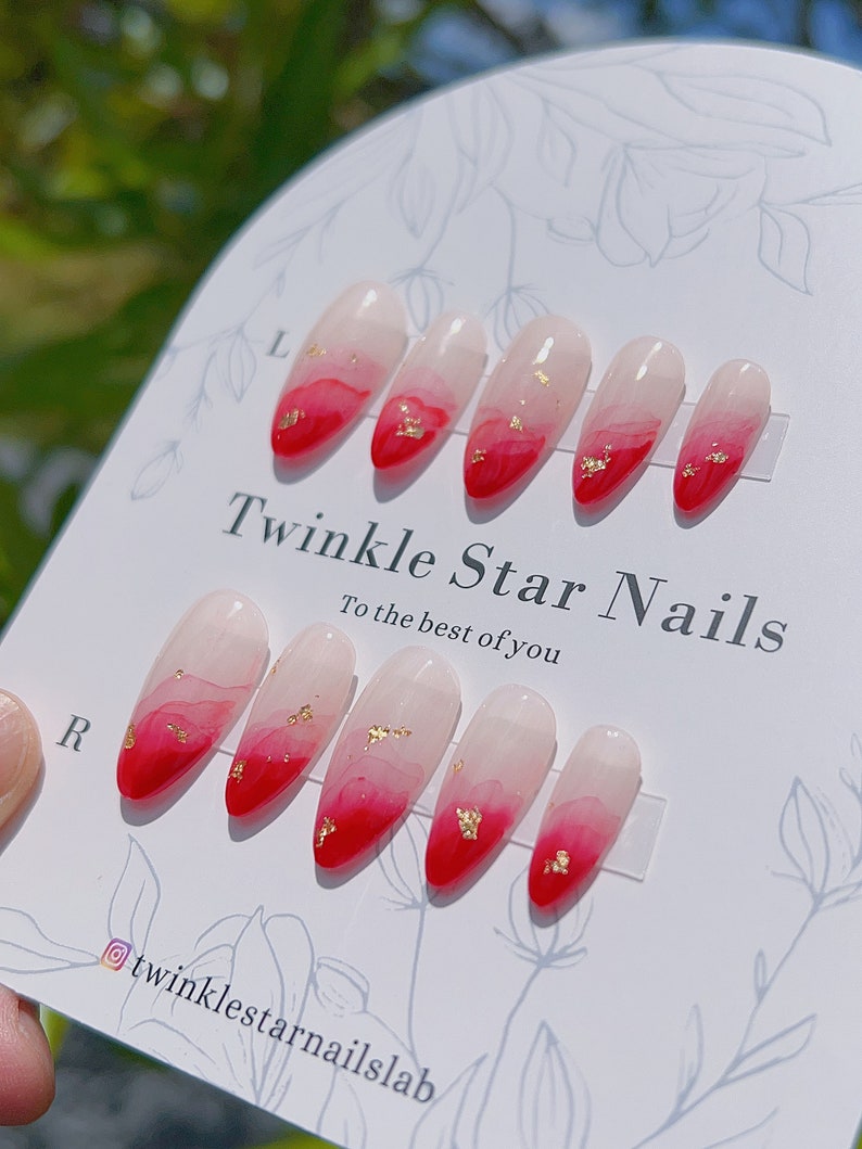 May include: Set of ten red and white ombre press-on nails with gold flakes. The nails are displayed on a white card with the text "Twinkle Star Nails, to the best of you" and the Instagram handle "@twinklestarnailslab".