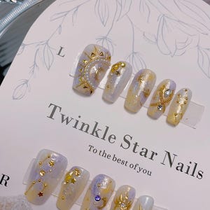 May include: A set of artificial nails with a celestial theme. The nails feature gold embellishments of stars, moons, and suns, set against a backdrop of pastel yellow, lavender, and white. The text "Twinkle Star Nails" is visible.