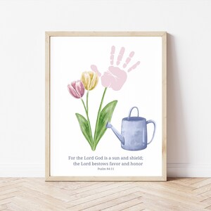 May include: Framed watercolour print with a light wood frame. The image shows a pink handprint, pink and yellow tulips, and a blue watering can. The text reads: "For the Lord God is a sun and shield; the Lord bestows favour and honour Psalm 84:11."