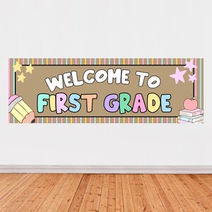 以下が含まれることがあります： 中央がベージュでストライプの縁取りが施された長方形のバナー。WELCOME TO FIRST GRADEというカラフルな文字が描かれています。黄色い鉛筆、ピンクの星、赤いリンゴ、積み重なった本のイラストがデザインに含まれています。