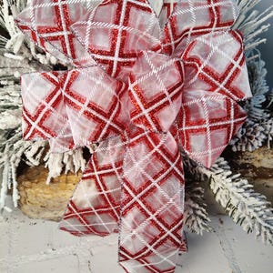 May include: A large decorative bow with a red and white plaid pattern. The bow is made of sheer ribbon with a glittery finish. The bow is likely for holiday or seasonal decor.