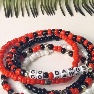 May include: Stack of beaded bracelets in red, black, and white. One bracelet spells out "GO DAWGS" with white letter beads. The bracelets have silver-colored accents and are arranged in a circular pattern.