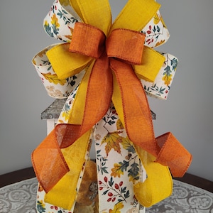 May include: A decorative bow featuring a mix of autumn colors. The bow has layers of yellow, orange, and patterned ribbons with fall leaf and floral designs. The bow is likely for seasonal decor.