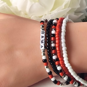 May include: A collection of beaded bracelets in red, black, white, and silver. One bracelet spells out "BAMA" in white letter beads. The bracelets are worn together, creating a layered look. The beads are small and round, with a mix of colors and textures.