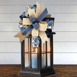 May include: A black metal lantern with glass panels containing a lit candle, topped with a decorative multi-layered bow. The bow features a combination of solid beige burlap, solid navy blue denim-textured fabric, and a floral print ribbon.