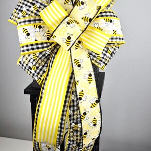 May include: A decorative bow featuring a mix of wired ribbons in yellow and white stripes, black and white buffalo check, and a cream-colored fabric printed with yellow bees and honeycomb patterns, tied into a large, multi-looped arrangement.