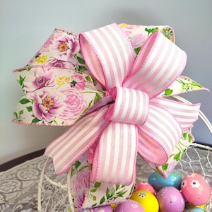 May include: A pink and white striped ribbon bow with a floral print ribbon underneath. The bow is sitting on top of a white wire basket filled with colorful Easter eggs.