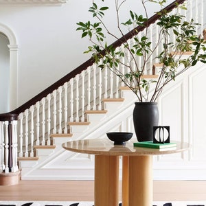 May include: A round wooden table with a marble top sits in a foyer with a white staircase and a black vase with green leafy branches. A black bowl and a green book are on the table. A black and white patterned rug is on the floor.