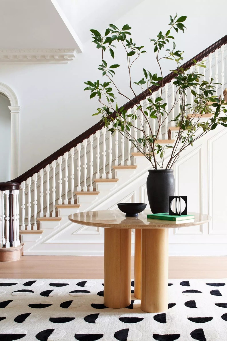 May include: A round wooden table with a marble top sits in a foyer with a white staircase and a black vase with green leafy branches. A black bowl and a green book are on the table. A black and white patterned rug is on the floor.