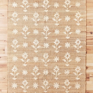 Puede incluir: Una alfombra rectangular de yute con un patr&oacute;n floral repetido. El color beige natural de la alfombra contrasta con el dise&ntilde;o floral blanquecino. Las flores est&aacute;n estilizadas con un tallo y hojas, y la alfombra est&aacute; colocada sobre un suelo de madera.