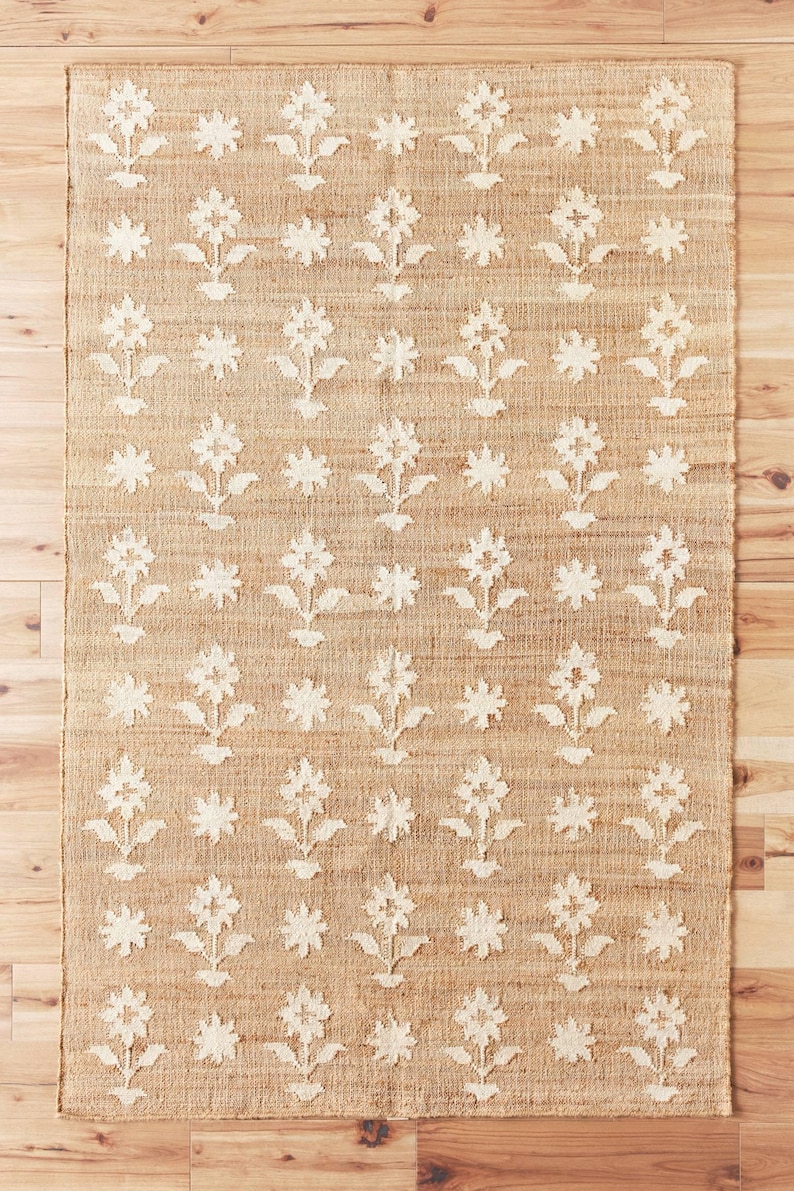 Puede incluir: Una alfombra rectangular de yute con un patr&oacute;n floral repetido. El color beige natural de la alfombra contrasta con el dise&ntilde;o floral blanquecino. Las flores est&aacute;n estilizadas con un tallo y hojas, y la alfombra est&aacute; colocada sobre un suelo de madera.
