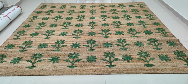 Op de afbeelding: Een vloerkleed van natuurlijke vezels met een geweven textuur en een herhalend patroon van groene bloemenmotieven.