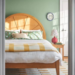 May include: A bedroom with a bed featuring a wooden headboard with a sunburst design. The bed is made with white linens and a yellow and white checkered blanket. A red lamp with a white and red patterned lampshade sits on a wooden nightstand next to the bed.