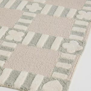 May include: A soft, patterned rug with a checkerboard design. The rug features squares of light pink and gray, with white stripes and floral and heart shapes. The rug is made of a plush material.