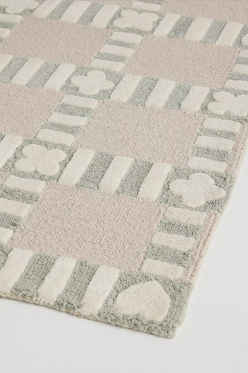 May include: A soft, patterned rug with a checkerboard design. The rug features squares of light pink and gray, with white stripes and floral and heart shapes. The rug is made of a plush material.