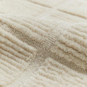 May include: Close-up of a cream-coloured rug with a textured, woven pattern. The rug has a soft, fluffy texture.