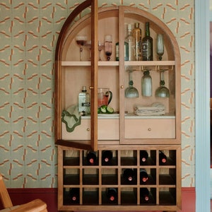 May include: A wooden bar cabinet with glass doors and shelves. The cabinet has a wine rack underneath and is decorated with a patterned wallpaper. The cabinet is open and shows various bottles of liquor, glasses, and bar tools.