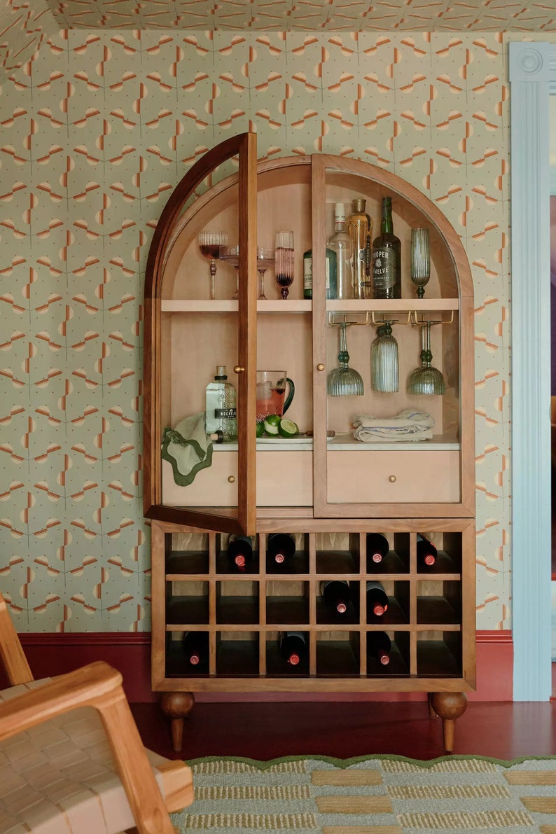 May include: A wooden bar cabinet with glass doors and shelves. The cabinet has a wine rack underneath and is decorated with a patterned wallpaper. The cabinet is open and shows various bottles of liquor, glasses, and bar tools.