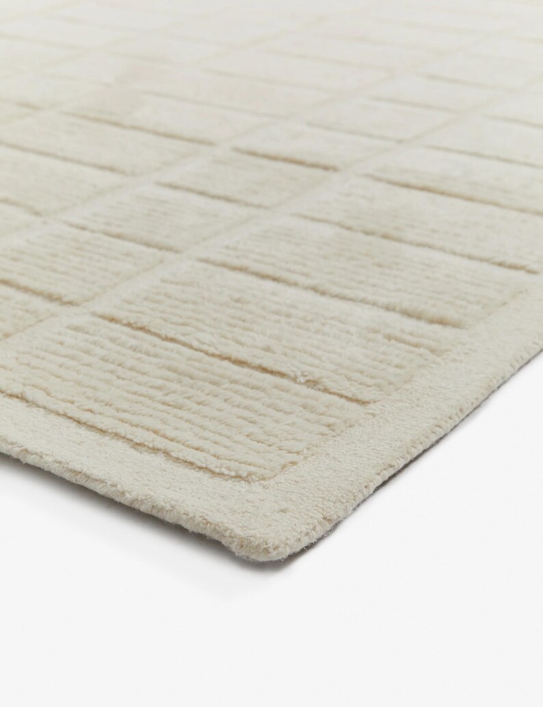 May include: A close-up of a cream-coloured rug with a textured, woven design. The rug has a subtle border around the edges.