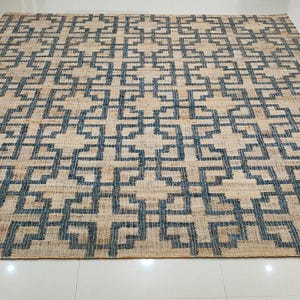 May include: A rectangular rug with a geometric pattern. The rug has a natural beige background with a repeating dark blue square and cross design. The rug is made of a woven material and is placed on a white tile floor.