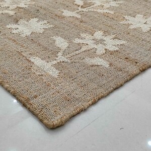 Puede incluir: Una alfombra rectangular de yute con un estampado floral. La alfombra es beige con dise&ntilde;os florales blancos. Se ve la esquina de la alfombra, mostrando la textura y el borde. La alfombra est&aacute; colocada sobre un suelo de baldosas blancas.