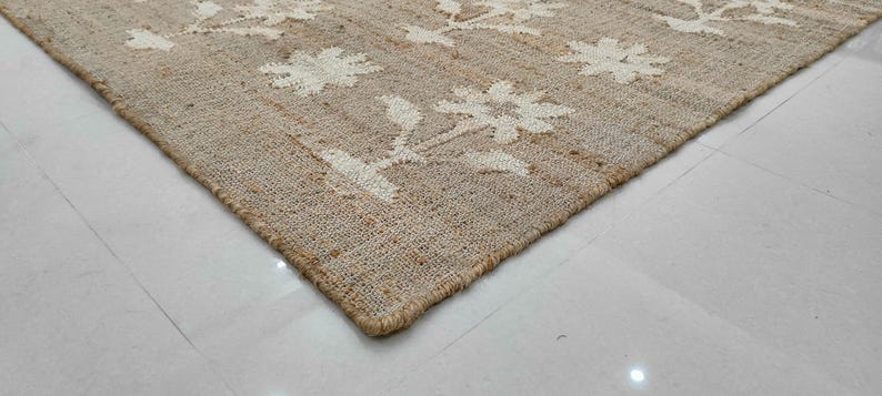 Puede incluir: Una alfombra rectangular de yute con un estampado floral. La alfombra es beige con dise&ntilde;os florales blancos. Se ve la esquina de la alfombra, mostrando la textura y el borde. La alfombra est&aacute; colocada sobre un suelo de baldosas blancas.