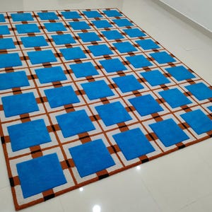 Op de afbeelding: Een rechthoekig tapijt met een herhalend patroon van blauwe vierkanten in een raster van bruine en zwarte lijnen op een witte achtergrond. Het tapijt is gemaakt van een zacht materiaal en is geschikt voor een woon- of slaapkamer.