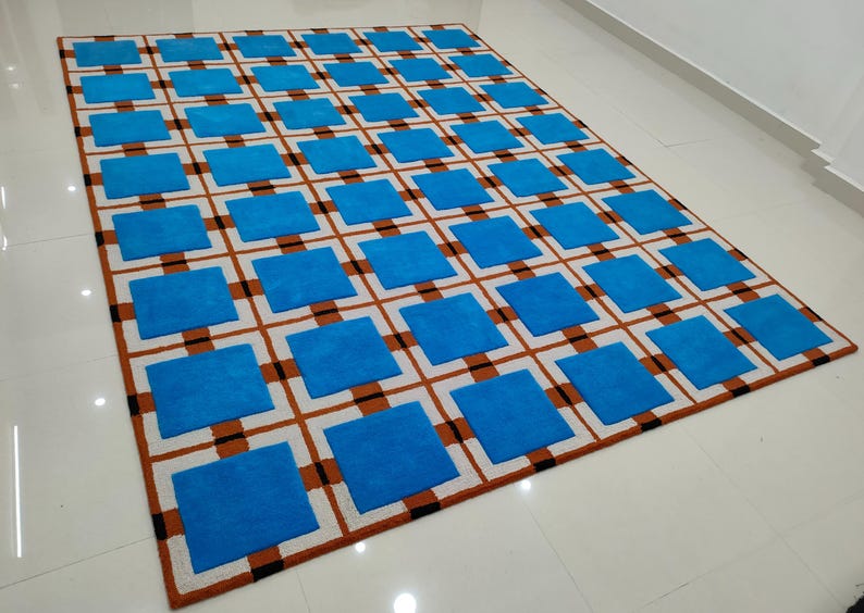 Op de afbeelding: Een rechthoekig tapijt met een herhalend patroon van blauwe vierkanten in een raster van bruine en zwarte lijnen op een witte achtergrond. Het tapijt is gemaakt van een zacht materiaal en is geschikt voor een woon- of slaapkamer.