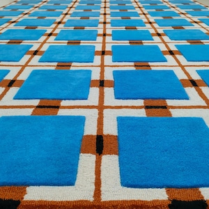 Op de afbeelding: Een groot tapijt met een herhalend patroon van blauwe vierkanten in een raster van witte, oranje en zwarte lijnen. Het tapijt heeft een getextureerd uiterlijk, wat wijst op een geweven of getuft constructie. Het ontwerp is geometrisch en modern.