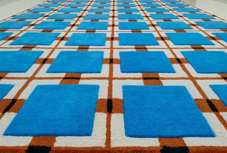 Op de afbeelding: Een groot tapijt met een herhalend patroon van blauwe vierkanten in een raster van witte, oranje en zwarte lijnen. Het tapijt heeft een getextureerd uiterlijk, wat wijst op een geweven of getuft constructie. Het ontwerp is geometrisch en modern.