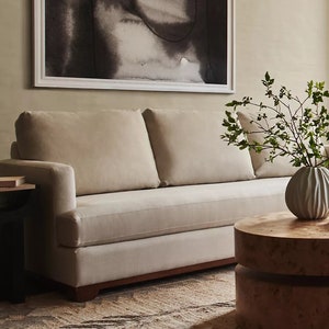 May include: A beige sofa with two cushions sits in a living room with a brown patterned rug. A wooden coffee table with a round top sits in front of the sofa. A vase with green branches sits on the table. A framed black and white abstract painting hangs on the wall behind the sofa.