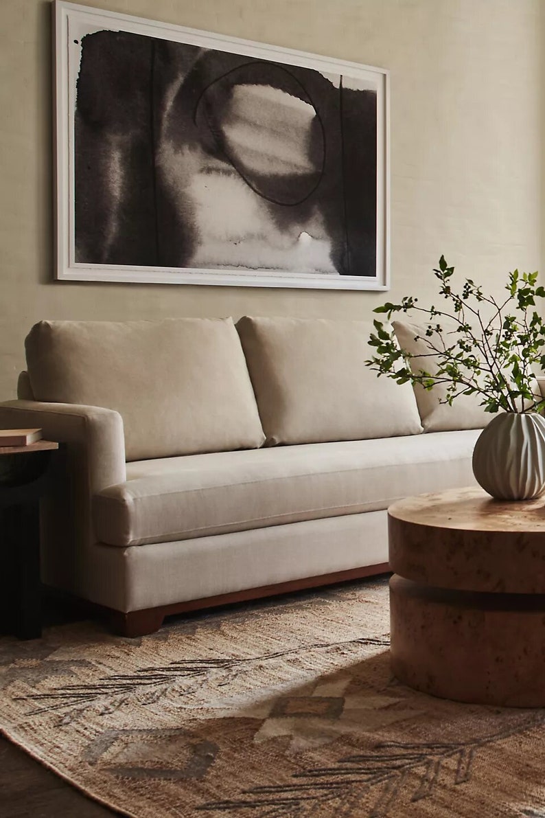 May include: A beige sofa with two cushions sits in a living room with a brown patterned rug. A wooden coffee table with a round top sits in front of the sofa. A vase with green branches sits on the table. A framed black and white abstract painting hangs on the wall behind the sofa.