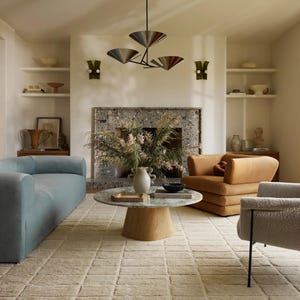 May include: A living room featuring a light blue sofa, a brown armchair, and a white rug with a geometric pattern. A coffee table with a marble top sits in the centre of the room. A fireplace with a stone surround is in the background. A chandelier with four lights hangs from the ceiling.