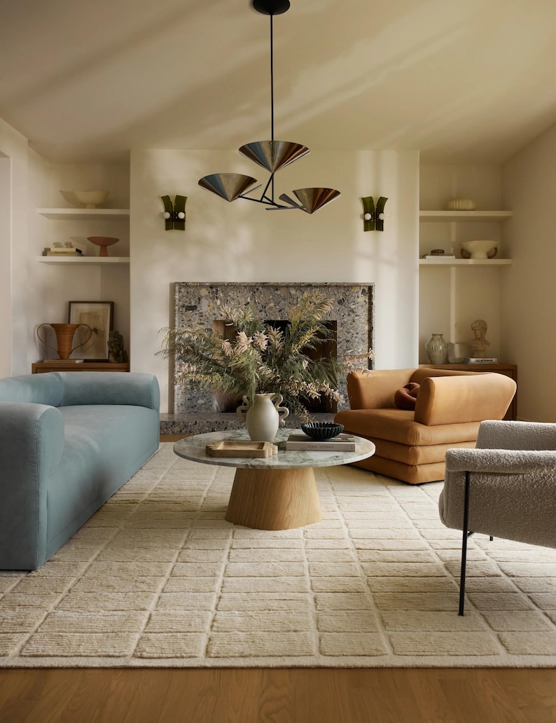 May include: A living room featuring a light blue sofa, a brown armchair, and a white rug with a geometric pattern. A coffee table with a marble top sits in the centre of the room. A fireplace with a stone surround is in the background. A chandelier with four lights hangs from the ceiling.
