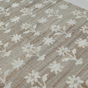 Puede incluir: Una alfombra rectangular beige con un patr&oacute;n floral blanco repetido. Las flores est&aacute;n estilizadas y dispuestas en filas sobre la superficie de la alfombra. La alfombra parece estar hecha de un material tejido, posiblemente yute o una fibra natural similar.