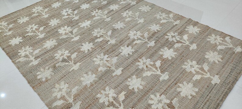 Puede incluir: Una alfombra rectangular beige con un patr&oacute;n floral blanco repetido. Las flores est&aacute;n estilizadas y dispuestas en filas sobre la superficie de la alfombra. La alfombra parece estar hecha de un material tejido, posiblemente yute o una fibra natural similar.