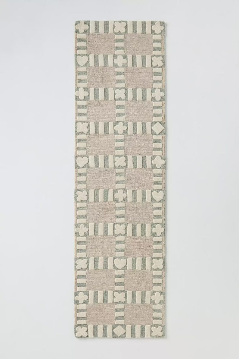 May include: A rectangular, patterned rug in a neutral color palette. The rug features a grid design with alternating squares of beige and gray, adorned with heart, diamond, and cross shapes. The rug is approximately 60 inches long.